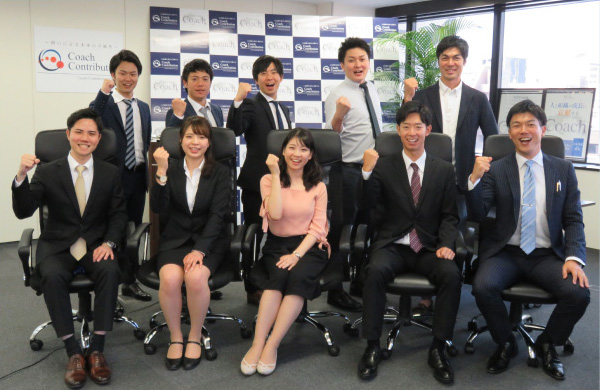写真:椅子に座っている3名の男性社員と2名の女性社員。その後ろには5名の男性社員が立っている。是認右手でガッツポーズをしている。